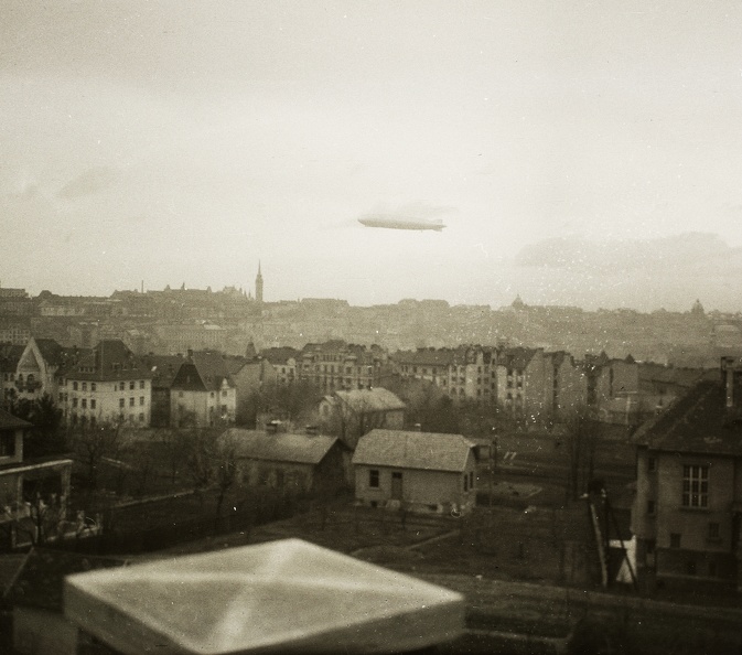 előtérben az Ügyész utca, szemben a Kék Golyó utca házsora, felette a budai Vár és a Graf Zeppelin léghajó.